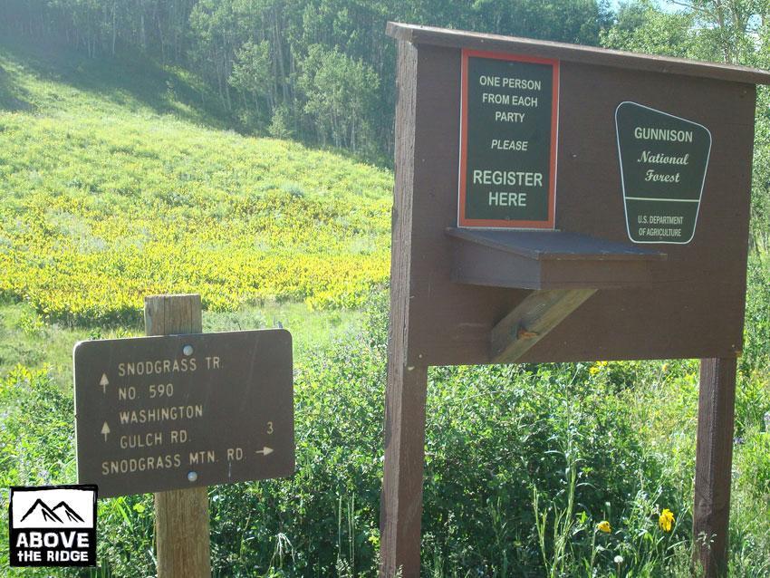 A wooden sign at the entrance of a trail in Gunnison National Forest. The sign states "One Person From Each Party Please Register Here" and features a green plaque with "Gunnison National Forest." Below, another sign gives directions to SnoDoggrass Trail No. 590 and indicates distances to Washington Gulch Road and SnoDoggrass Mountain Road. The background shows a vibrant landscape with green hills and scattered wildflowers. Snodgrass mountain bike trail.