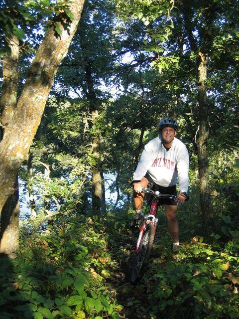 A person riding a mountain bike on a narrow, wooded trail surrounded by greenery and trees, with sunlight filtering through the leaves. There is a body of water visible in the background. Lake Metigoshe State Park mountain bike trail.