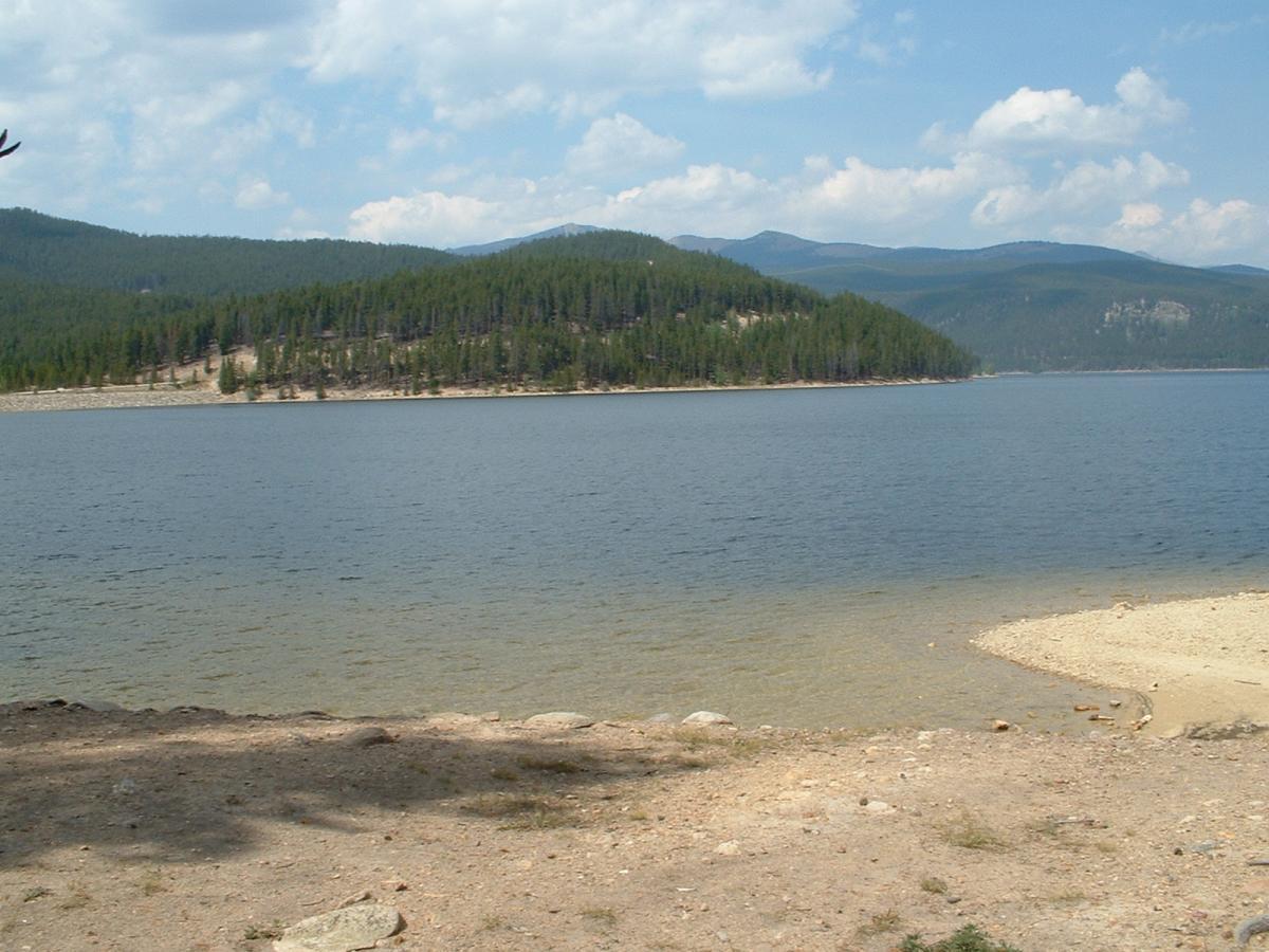 A serene lakeside view featuring calm waters surrounded by rolling hills and dense pine forests under a partly cloudy blue sky. The sandy shore in the foreground gently slopes into the lake, creating a peaceful natural setting. Turquoise Lake mountain bike trail.