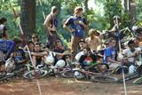 A group of young people, some wearing helmets and riding gear, gathered around bicycles in a wooded area. They are sitting and standing together, engaged in conversation and enjoying their time outdoors, with trees and dirt paths visible in the background. Georgia International Horse Park mountain bike trail.