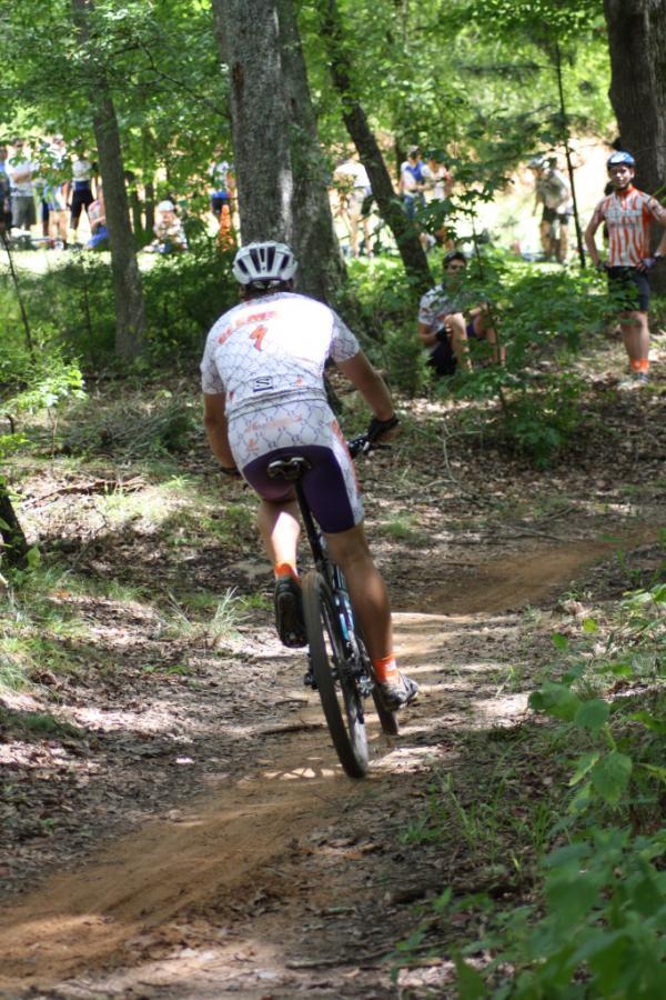 A cyclist wearing a white and purple racing jersey navigates a winding dirt trail in a wooded area, with spectators in the background watching the race. The scene conveys a sense of action and excitement in a natural setting. Georgia International Horse Park mountain bike trail.