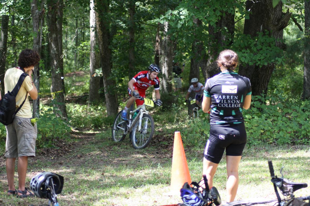 A mountain biking event taking place in a wooded area. In the foreground, a participant in a red jersey with the number 33 is navigating a trail. Two spectators, one taking photos and another wearing a jersey from Warren Wilson College, observe the race, while bicycles are resting on the ground nearby. The scene is filled with greenery, showcasing a sunny day outdoors. Georgia International Horse Park mountain bike trail.