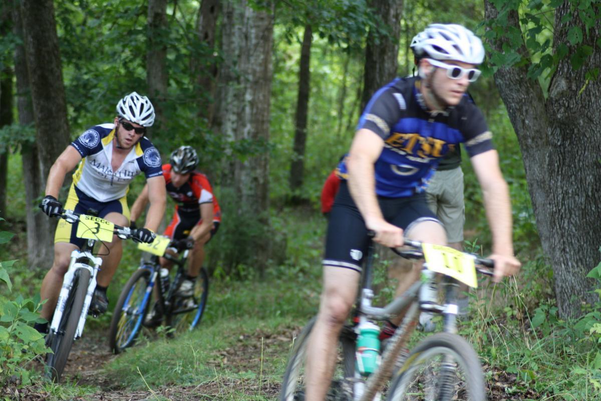 A group of mountain bikers navigating a wooded trail. The foreground shows two cyclists in brightly colored jerseys, one wearing sunglasses and the other slightly blurred as they ride downhill. The background features trees and greenery, indicating a natural outdoor setting. A third biker is partially visible in the background, adding to the dynamic of the race. Georgia International Horse Park mountain bike trail.