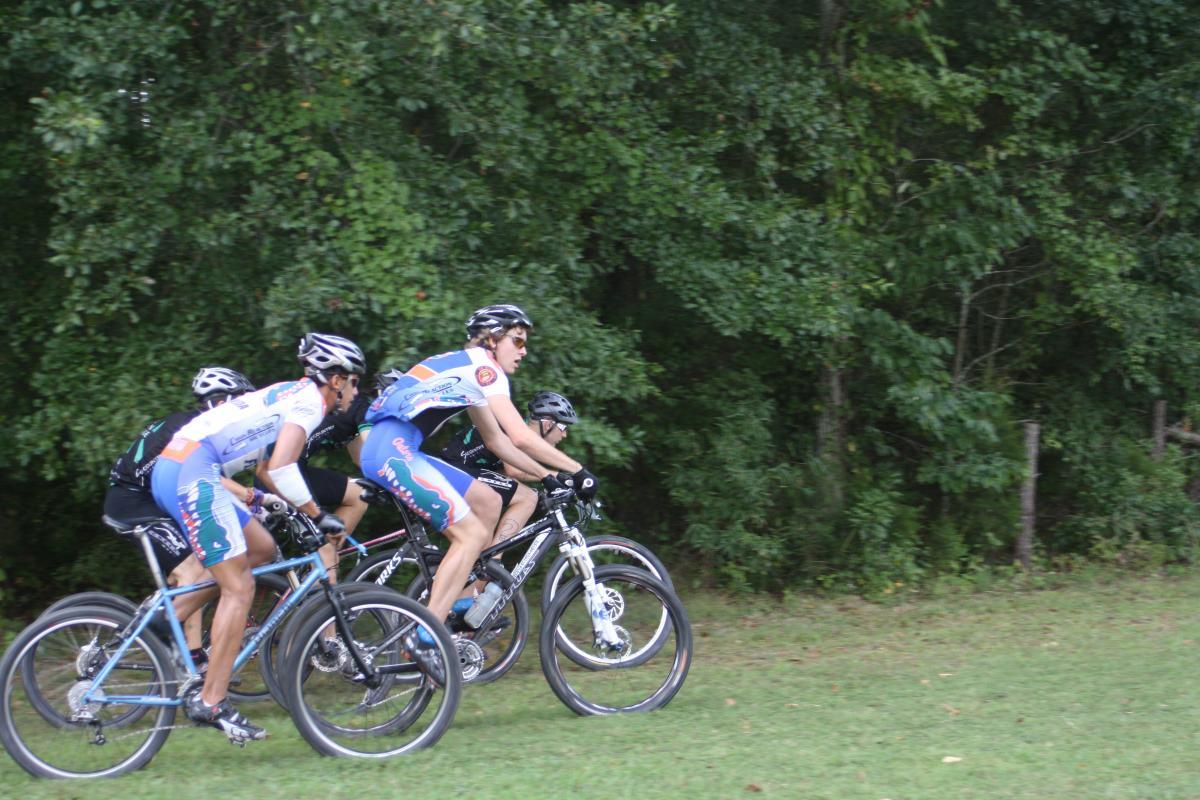 Three cyclists in athletic clothing are riding their bikes on a grassy trail, surrounded by trees. The cyclists are focused and moving in formation, showcasing a competitive atmosphere. Two cyclists are wearing jerseys with colorful designs, while the third is dressed in black. Georgia International Horse Park mountain bike trail.