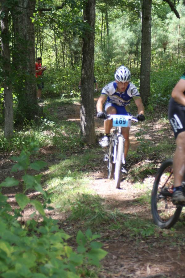 A cyclist in a blue and white racing jersey is riding a mountain bike along a narrow trail in a wooded area. The cyclist is wearing a helmet and has a race number visible on their jersey. In the background, another person stands near the trail, observing the event. The scene is surrounded by greenery and trees, indicating a natural outdoor setting. Georgia International Horse Park mountain bike trail.