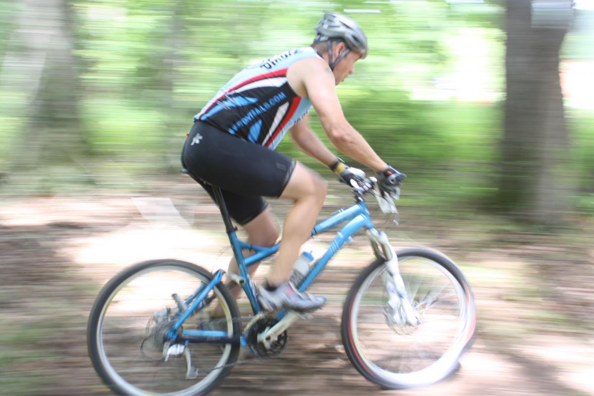 A motion blur image of a cyclist in a racing outfit riding a blue mountain bike on a dirt trail surrounded by trees. The cyclist is captured mid-motion, emphasizing speed and agility. Georgia International Horse Park mountain bike trail.