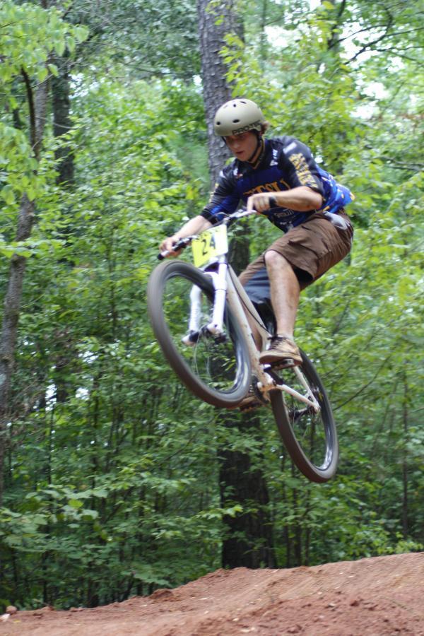 A mountain biker in mid-air performing a jump over a dirt ramp, surrounded by lush green trees. The rider is wearing a helmet and protective gear, with a number plate visible on their bike. Georgia International Horse Park mountain bike trail.
