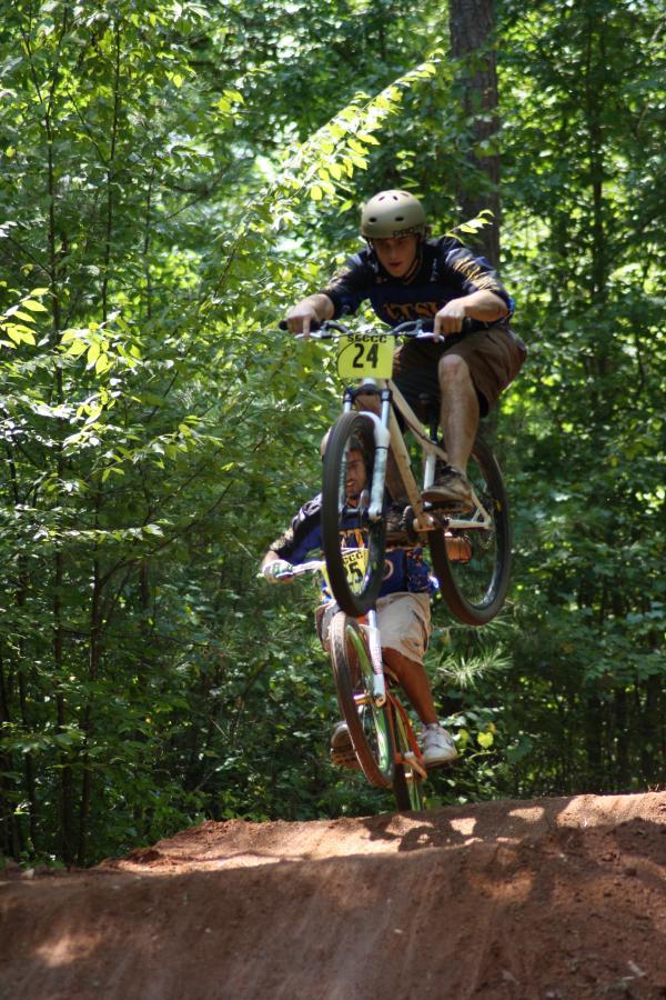 Two BMX riders performing tricks on a dirt jump in a forested area. One rider is airborne while the other is positioned below, both wearing helmets and casual clothing. The surrounding greenery suggests a lively outdoor environment. Georgia International Horse Park mountain bike trail.