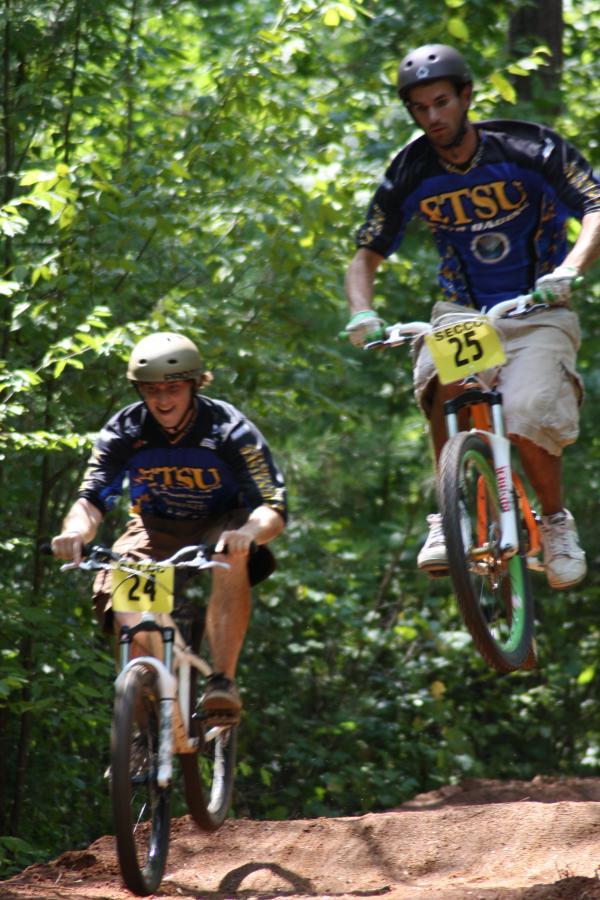 Two mountain bikers participating in a race on a dirt trail surrounded by trees. One rider, wearing a blue and black jersey with the number 24, is pedaling, while the other, in a similar jersey with the number 25, is airborne on his bike, having just jumped off a small hill. Both riders are wearing helmets for safety. Georgia International Horse Park mountain bike trail.