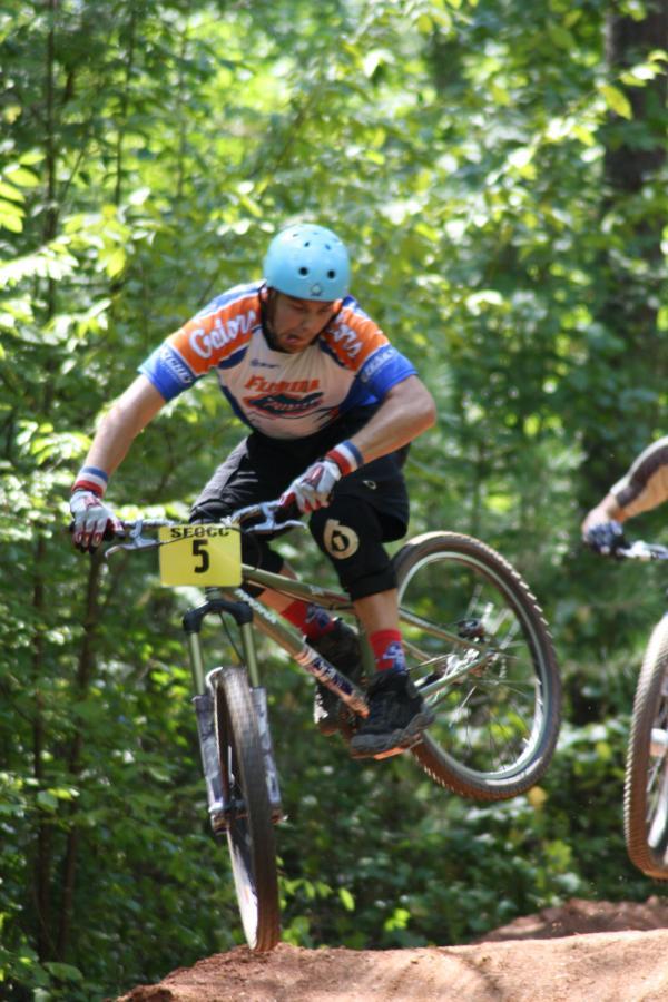 A mountain biker in a blue helmet and a colorful jersey performs a jump on a dirt trail surrounded by green trees. The rider is wearing black shorts and colorful socks, with the bike's number plate visible on the front. Another biker can be seen in the background, also navigating the trail. Georgia International Horse Park mountain bike trail.