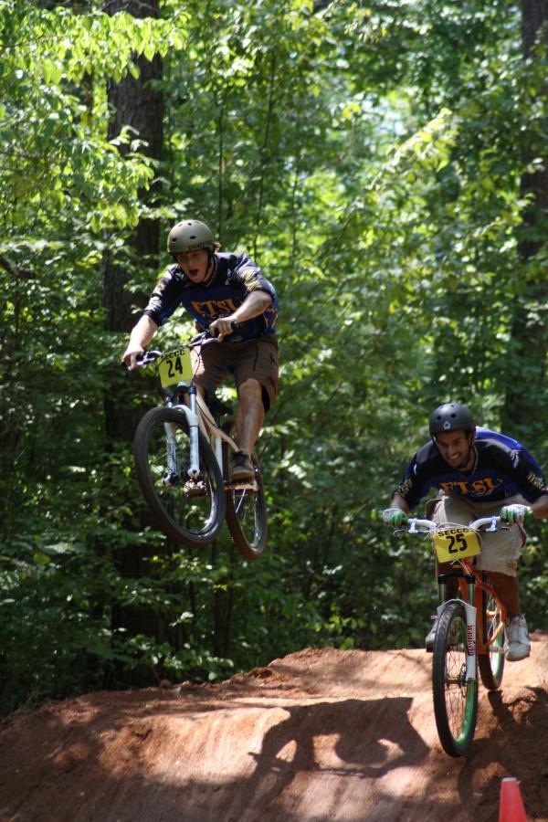Two mountain bikers performing jumps on a dirt track surrounded by trees. The biker on the left is airborne, wearing a helmet and a black jersey with the number 24. The biker on the right, also in a helmet and a jersey with the number 25, is landing after a jump. Sunlight filters through the leaves, creating a vibrant outdoor scene. Georgia International Horse Park mountain bike trail.