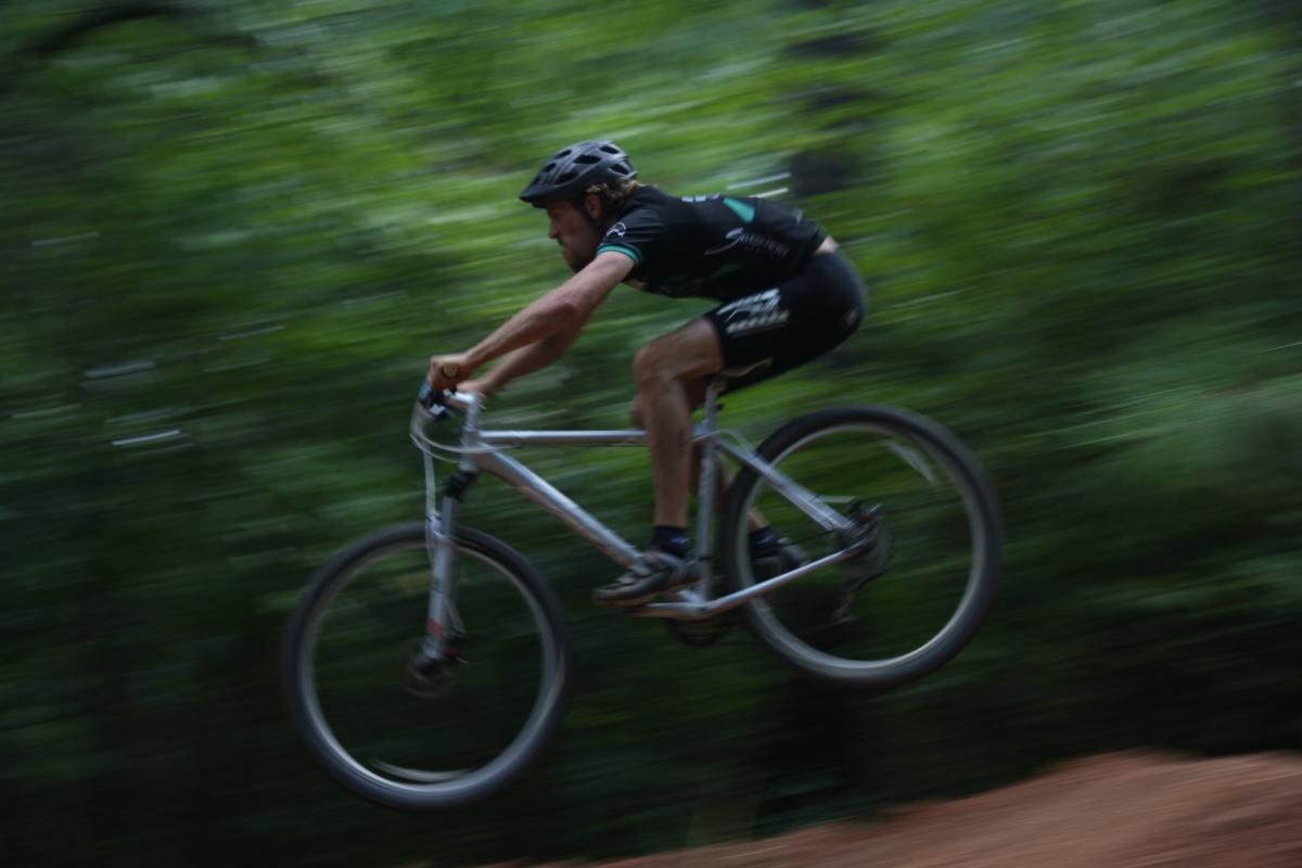 A mountain biker jumps into the air while riding on a dirt trail surrounded by greenery, captured in motion with a blurred background to emphasize speed and action. Georgia International Horse Park mountain bike trail.
