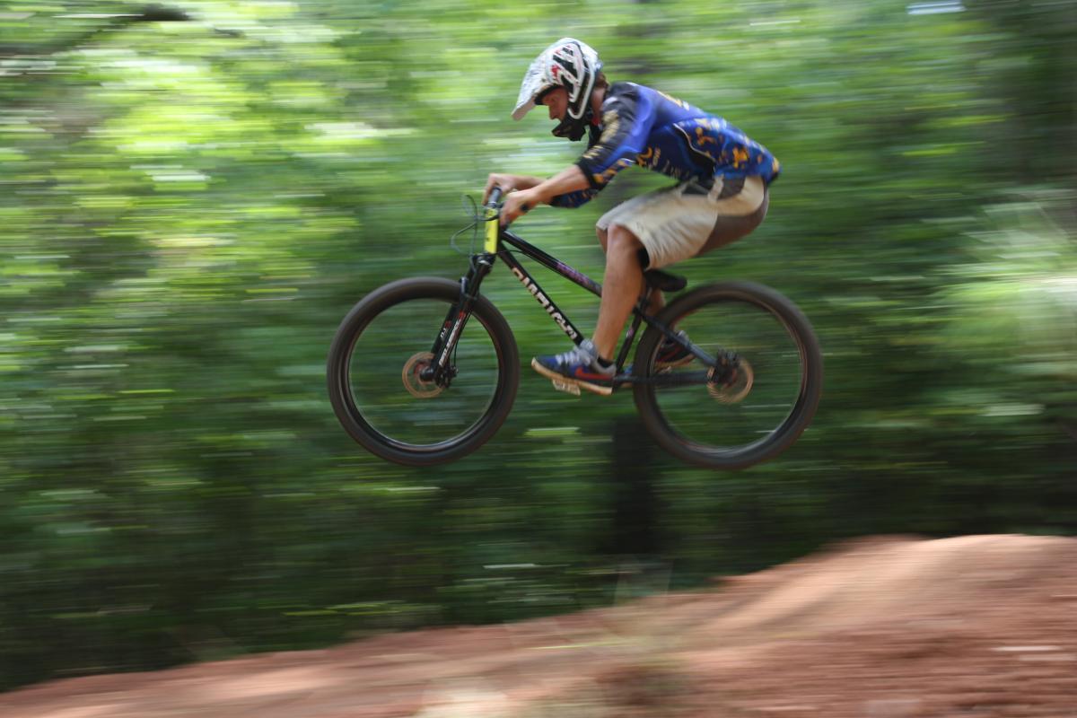 A cyclist performing a jump over a dirt ramp in a forested area, captured in motion with a blurred background to convey speed. The cyclist is wearing a helmet and a blue jersey, with their bike tilted as they ascend into the air. Georgia International Horse Park mountain bike trail.