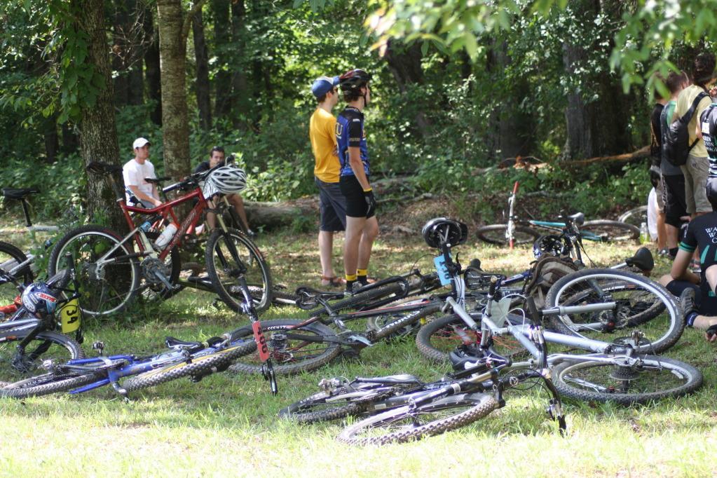 A group of mountain bikers resting in a wooded area, surrounded by several parked bicycles scattered on the grass. Some riders are standing and chatting, while others are seated nearby. The scene is lush with greenery, indicating a warm, sunny day outdoors. Georgia International Horse Park mountain bike trail.