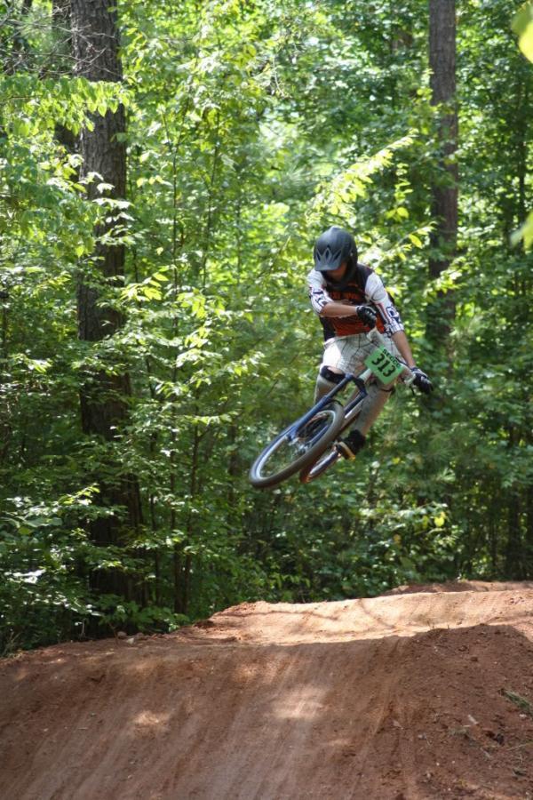 A young cyclist performs a jump on a dirt trail surrounded by lush green trees. The rider is wearing protective gear, including a helmet and a jersey with a race number. Sunlight filters through the foliage, creating a vibrant outdoor atmosphere. Georgia International Horse Park mountain bike trail.