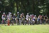 A group of cyclists lined up on their mountain bikes in a grassy area, preparing for a race. They are wearing athletic gear and helmets, with trees in the background indicating an outdoor setting. Georgia International Horse Park mountain bike trail.