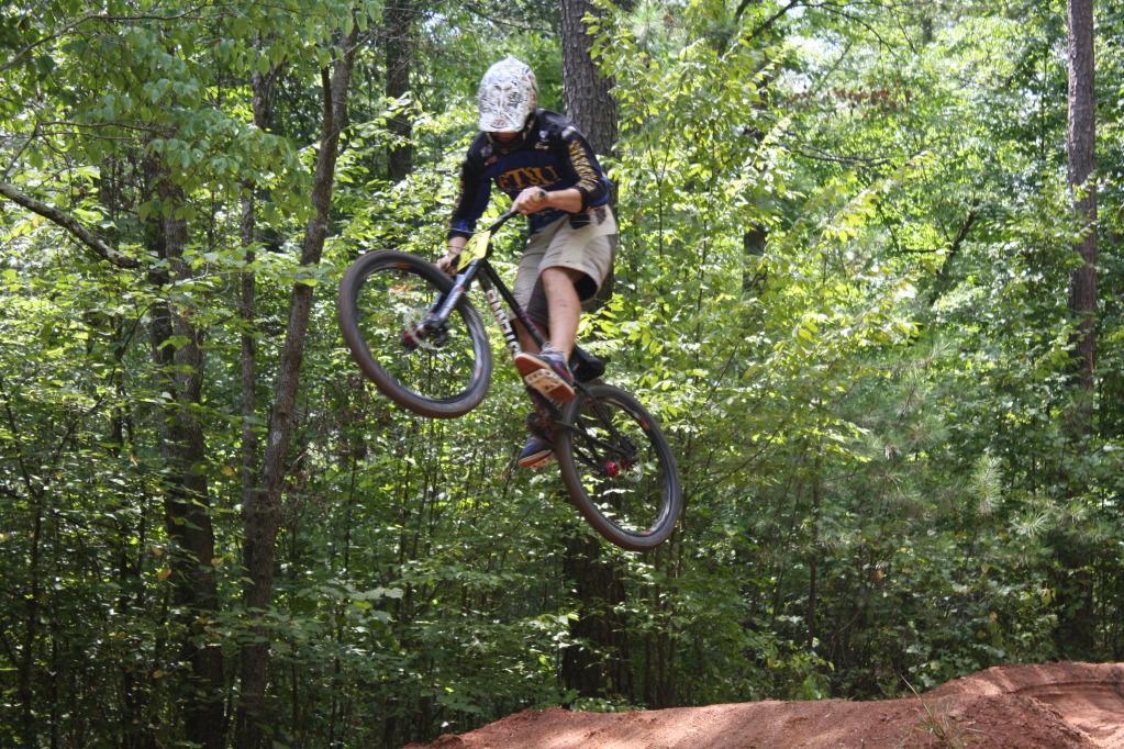 A person in a black shirt and beige shorts is performing a jump on a mountain bike in a wooded area. The cyclist is airborne, with both wheels off the ground, surrounded by lush green trees. Georgia International Horse Park mountain bike trail.