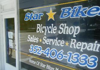 A close-up view of a storefront window displaying the sign for "Star Bikes," a bicycle shop that offers sales, service, and repair. The window includes contact information (phone number) and highlights its offerings. The scene captures a sunny day outside.