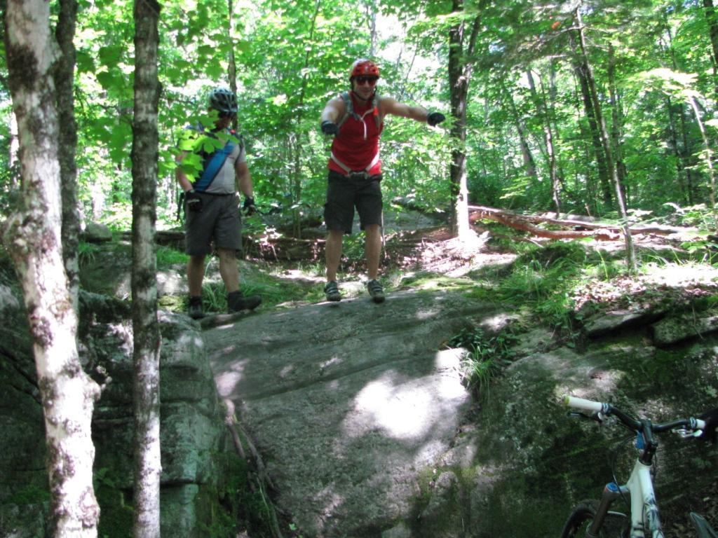 Two mountain bikers navigating a rocky trail in a green, wooded area. One biker, wearing a red sleeveless jersey and helmet, is demonstrating a pose while standing on a rock. The other biker, in a blue shirt and shorts, is watching nearby. A bicycle can be seen on the right side of the image. Sunlight filters through the tree canopy, illuminating the scene. Buckwallow mountain bike trail.