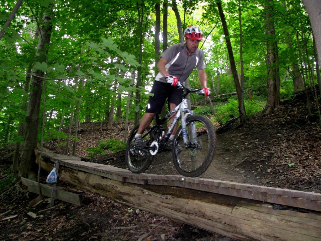 A man riding a mountain bike on a narrow wooden bridge in a lush green forest. The cyclist is wearing a helmet and gloves, and is focused on navigating the uneven terrain. Surrounding him are tall trees and thick foliage, indicating a vibrant outdoor setting. Don Valley mountain bike trail.