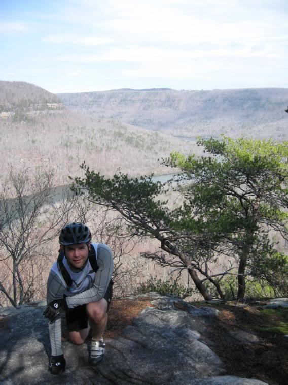 A person wearing a helmet and cycling gear kneels on a rocky outcrop, overlooking a valley with trees and a river below. The landscape features rolling hills and a clear sky in the background. Raccoon Mountain Trail Network mountain bike trail.