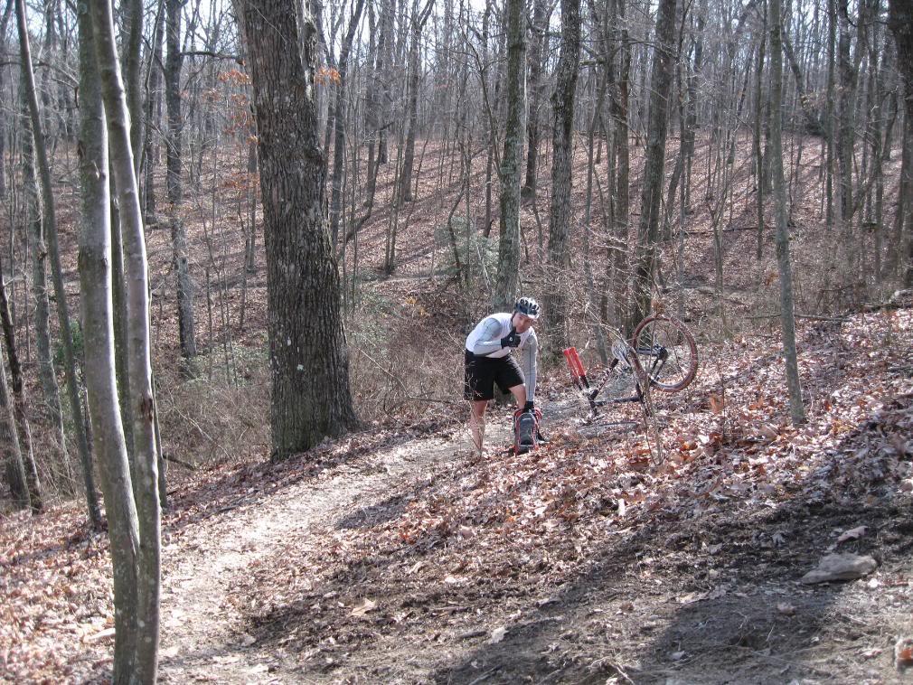 A mountain biker crouches next to his bike on a dirt path in a wooded area, surrounded by trees and fallen leaves. The bike is positioned with its front wheel in the air, indicating that it may have trouble. The sun is shining through the branches, casting soft shadows on the ground. The biker is wearing a helmet and a light, long-sleeve jersey, looking down as he examines his equipment. Raccoon Mountain Trail Network mountain bike trail.