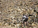 A mountain biker navigating through a rocky terrain with large boulders in the background on a bright, sunny day. Raccoon Mountain Trail Network mountain bike trail.