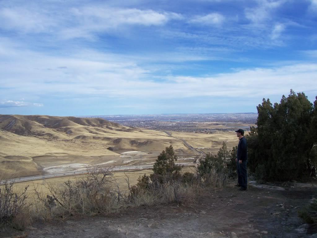 A person stands on a rocky overlook, gazing at a vast landscape of rolling hills and valleys under a blue sky with wispy clouds. Sparse vegetation and shrubs are visible in the foreground, with a winding road and distant cityscape in the background. Red Rocks / Dakota Ridge mountain bike trail.
