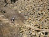 A person riding a mountain bike along a narrow wooden path, surrounded by rocky terrain and sparse vegetation. Raccoon Mountain Trail Network mountain bike trail.
