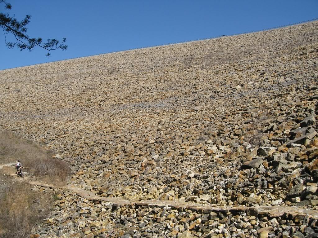 A rocky slope of a dam or hillside with a blue sky above; a narrow wooden path runs along the side, and a cyclist can be seen near the bottom of the image, navigating the rocky terrain. Raccoon Mountain Trail Network mountain bike trail.