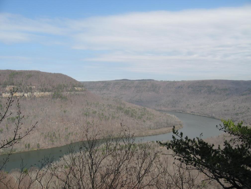 A panoramic view of a river winding through a wooded landscape, with bare trees and rolling hills under a cloudy sky. The scene captures the natural beauty of the area, showcasing the rugged terrain and the river's curves. Raccoon Mountain Trail Network mountain bike trail.