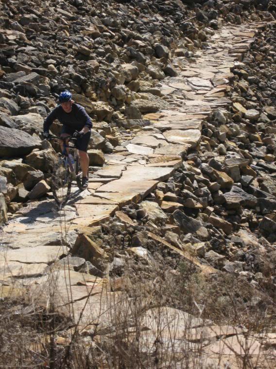 A cyclist navigating a rocky path surrounded by uneven terrain, with large stones and dried vegetation in the background. The cyclist is wearing a helmet and riding a mountain bike along a narrow, stony trail. Raccoon Mountain Trail Network mountain bike trail.