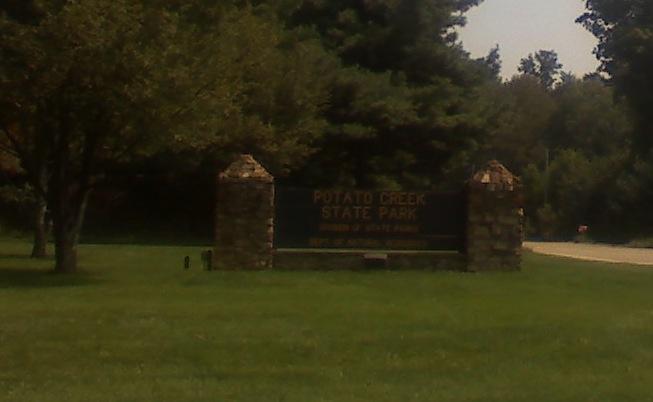 Sign for Potato Creek State Park, featuring stone pillars and surrounded by green grass and trees. The sign indicates the park's name and its designation as a state park. Potato Creek State Park mountain bike trail.