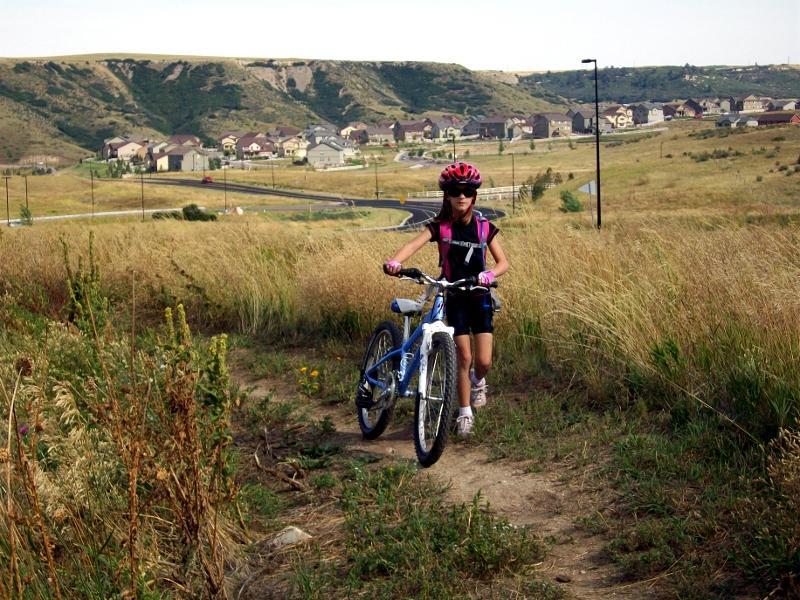 A young girl wearing a helmet and sunglasses is walking her mountain bike along a grassy path, with a scenic backdrop of rolling hills and a residential area in the distance. Rhyolite Bike Park mountain bike trail.