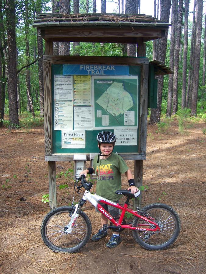 A young boy wearing a helmet and a green shirt stands next to a trail map sign for the Firebreak Trail in a wooded area. He smiles while holding onto a red and white bicycle, surrounded by tall trees and pine needles on the ground. Harbison State Forest mountain bike trail.
