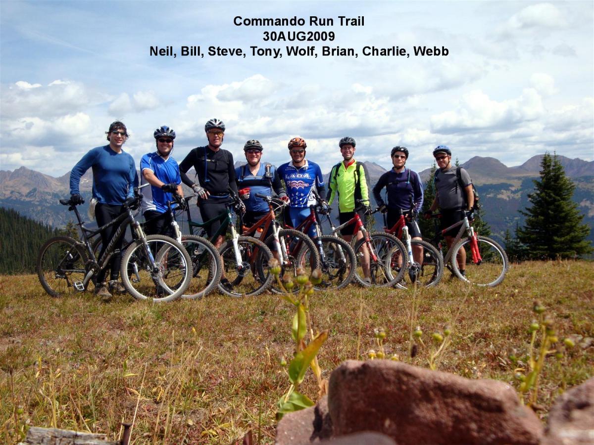 A group of eight mountain bikers stands on a grassy area with mountains in the background. They are dressed in cycling attire and are each holding their bicycles. The sky is partly cloudy, and a few trees are visible nearby. Commando Run mountain bike trail.