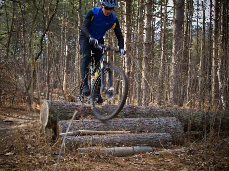 A mountain biker jumps over a series of logs on a forest trail, surrounded by tall trees and underbrush, showcasing a dynamic moment in outdoor cycling. Carvin's Cove Trail system mountain bike trail.