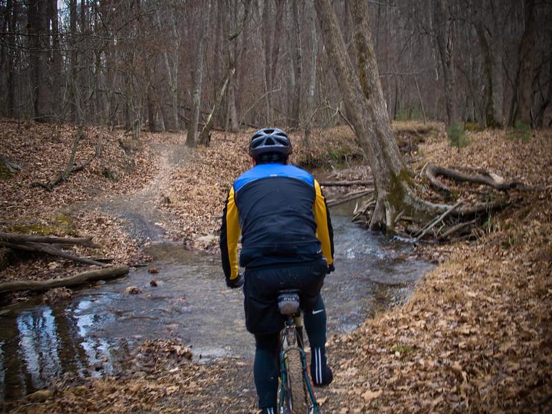 A cyclist navigating a muddy trail through a forested area, with fallen leaves covering the ground and a small stream flowing alongside the path. The scene is set in a wintry environment, showcasing bare trees and a winding trail ahead. Carvin's Cove Trail system mountain bike trail.