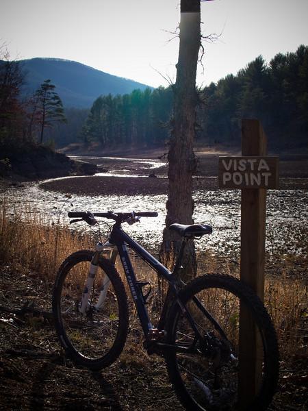 A mountain bike rests against a tree near a sign that reads "Vista Point," overlooking a dry riverbed surrounded by lush green trees and distant mountains under a bright sky. Carvin's Cove Trail system mountain bike trail.