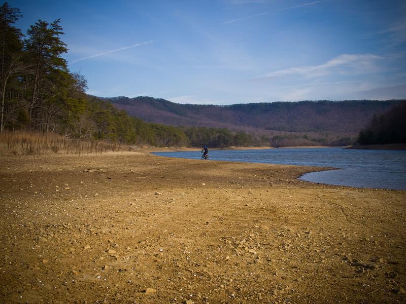 A sandy shore with sparse vegetation lines a calm lake, bordered by rolling hills in the background. A person is seen riding a bicycle near the water's edge under a clear blue sky. The scene is tranquil, showcasing the natural beauty of the landscape. Carvin's Cove Trail system mountain bike trail.