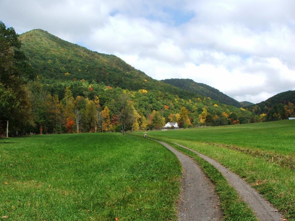 A scenic view of a grassy path winding through a vibrant autumn landscape, featuring colorful trees and rolling hills under a partly cloudy sky. In the distance, a charming farmhouse is visible among the trees. Virginia Creeper - White Top To Damascus mountain bike trail.