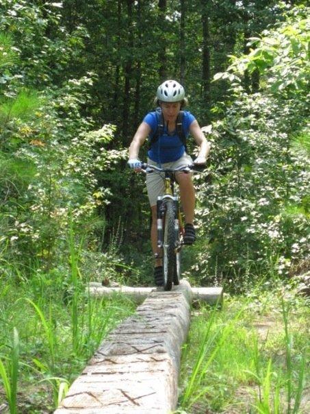 A person riding a mountain bike on a narrow log balance beam, surrounded by a lush green forest. The cyclist is wearing a blue shirt, shorts, and a helmet, showcasing an adventurous spirit in a natural setting. Noxubee Crest mountain bike trail.