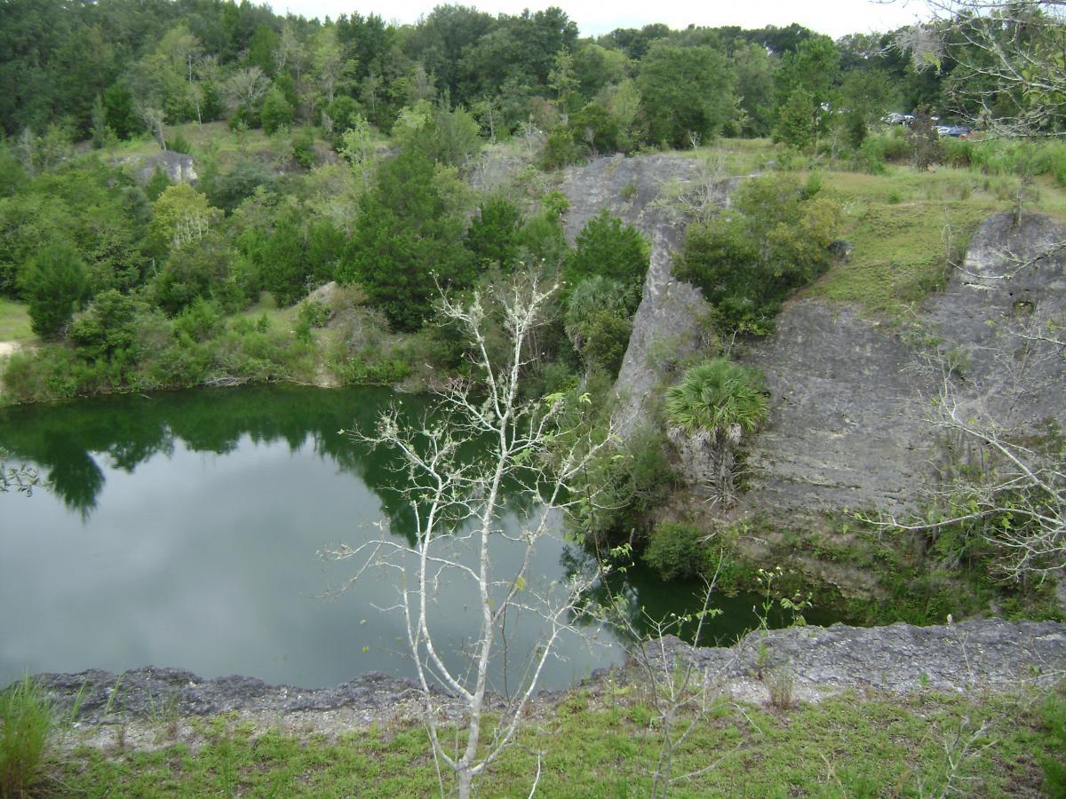 A tranquil view of a green pond surrounded by rocky cliffs and lush vegetation. The water reflects the surrounding trees, creating a peaceful natural scene. Haile's Trails mountain bike trail.