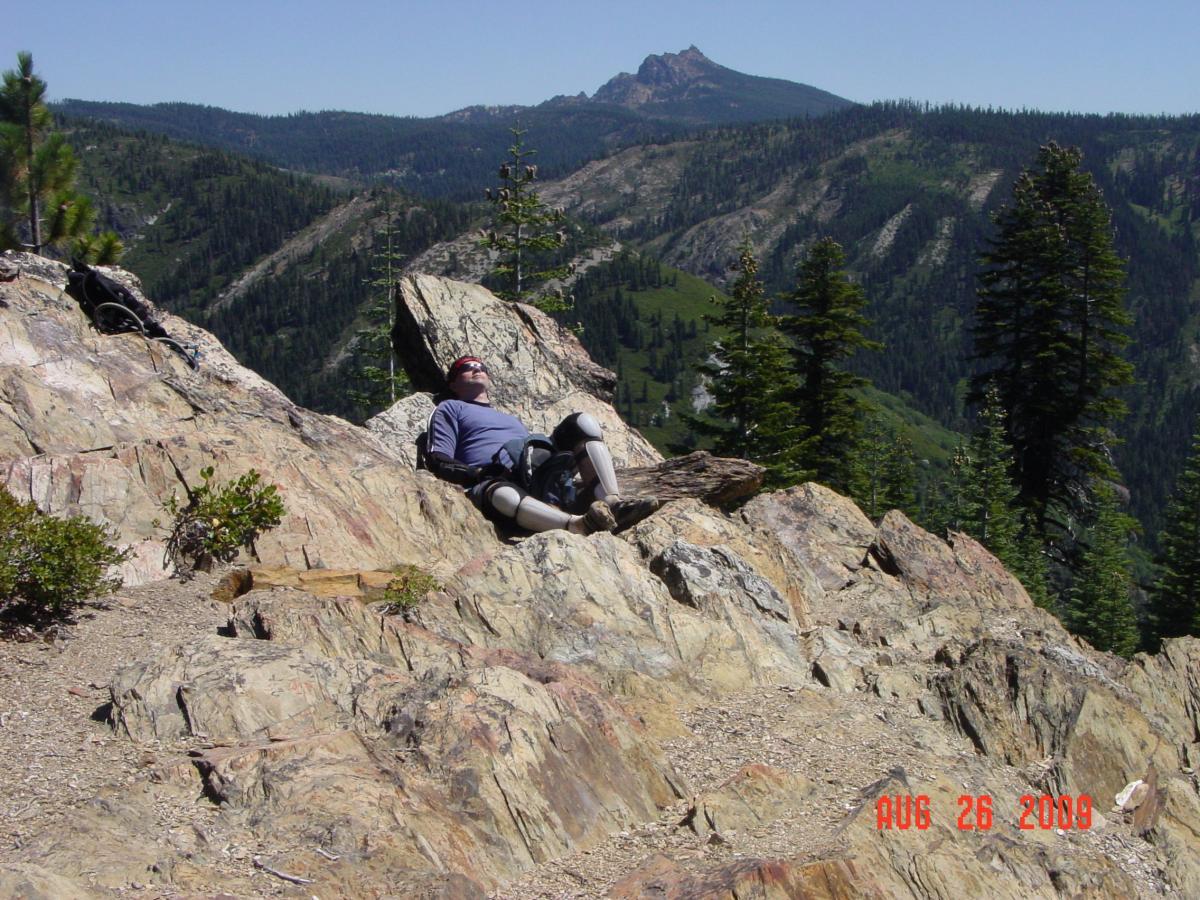 A person relaxing on a large rock in a mountainous area, surrounded by trees and a panoramic view of hills and valleys under a clear blue sky. Big Boulder Trail mountain bike trail.