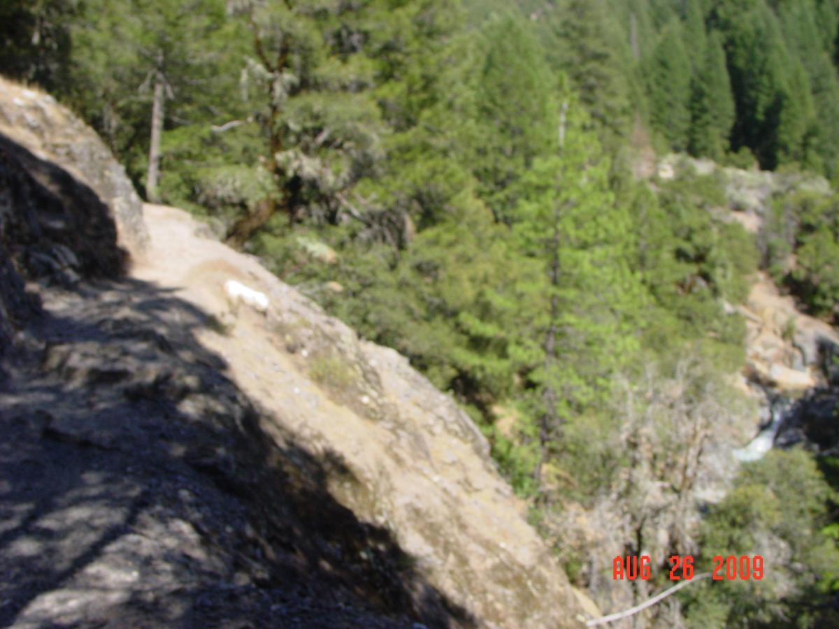 A rocky hiking trail curves along a steep cliffside, surrounded by dense green trees and vegetation. A river can be seen at the bottom of the canyon. The image is bright and taken during daylight. Downieville Downhill mountain bike trail.