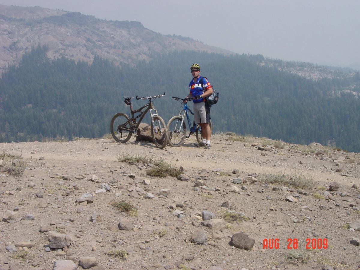 A cyclist stands on a rocky trail with two mountain bikes beside him. The background features a hazy mountain landscape with a dense forest of pine trees. The cyclist is wearing a helmet and a blue jersey with red accents, and he appears to be taking a break. The image is dated August 28, 2009. Hole In The Ground mountain bike trail.