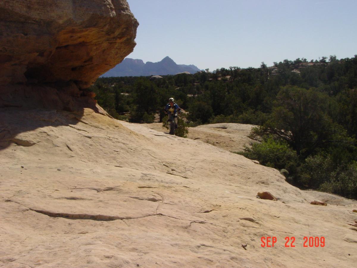 A rocky landscape with a person on a dirt bike near a large boulder. The background features distant mountains and a clear blue sky, with trees and shrubs scattered across the terrain. The image is dated September 22, 2009. Gooseberry Mesa mountain bike trail.