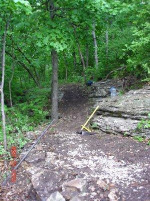 A narrow dirt path winding through a lush green forest, flanked by rocky outcrops and trees. A yellow tool is visible on the ground, along with some scattered rocks and a water bottle near the edge of the path. Swope Park Trail mountain bike trail.