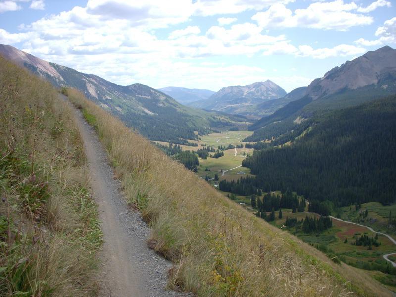 A scenic mountain trail winding along a hillside, overlooking a lush valley surrounded by peaks and forests under a partly cloudy sky. Trail 401 mountain bike trail.