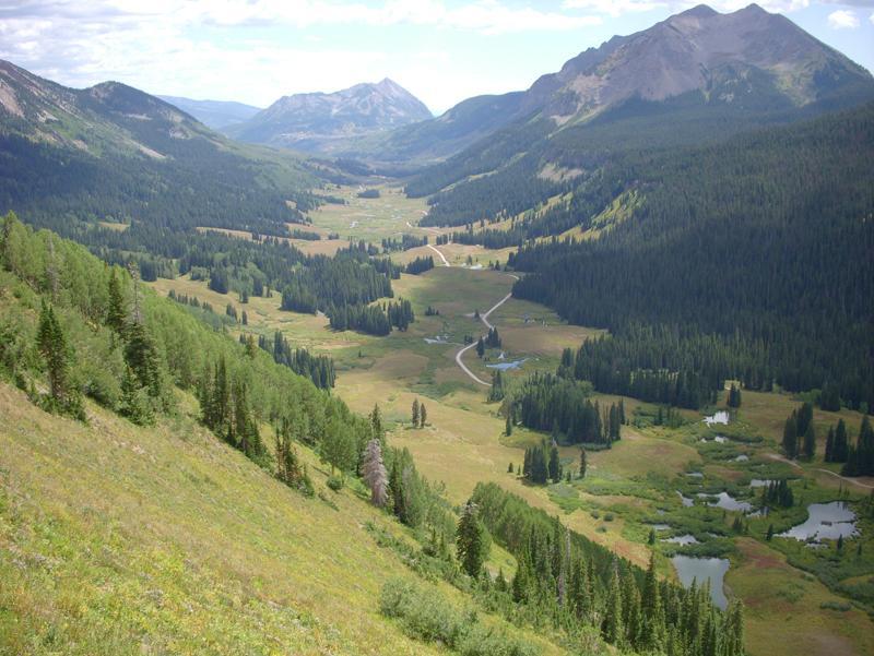 A panoramic view of a lush green valley surrounded by mountains. The landscape features rolling hills with patches of trees, winding roads, and scattered lakes, under a partly cloudy blue sky. Trail 401 mountain bike trail.